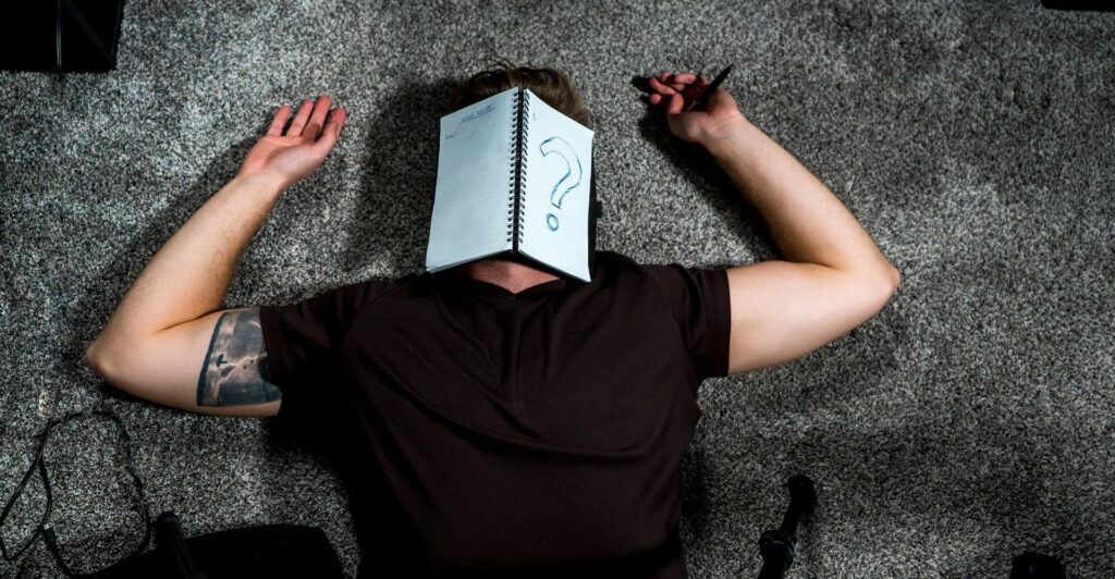 A man lies on his back with a notebook over his face. A large question mark is drawn on one page.