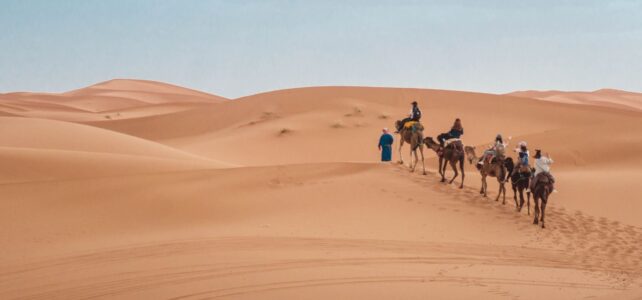 Half a dozen people riding camels are led through a barren desert.